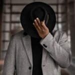 Elegant photo of a man in a formal coat and fedora hat posing indoors.
